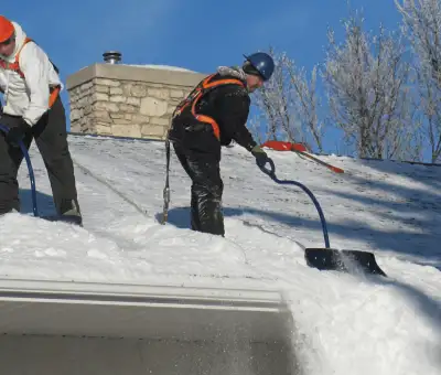 Professional technicians performing roof snow removal with safety harnesses and shovels in Fargo, ND